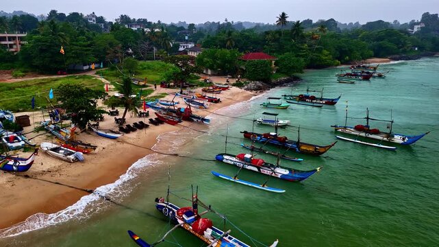 Aerial view of traditional outrigger fishing boats anchored along the sandy coastline with lush tropical vegetation in the background, Sri Lanka.