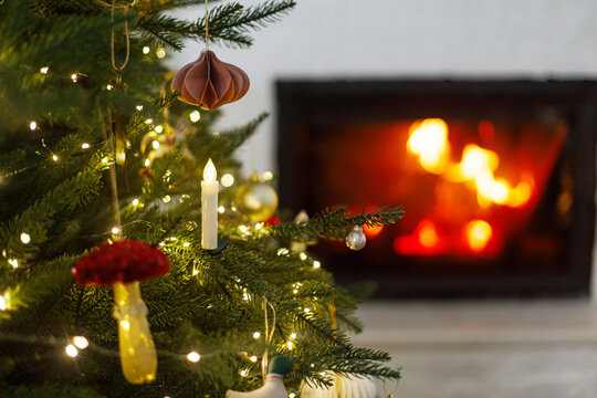Christmas tree with beautiful baubles, garland lights and paper ornaments. Warm fire burning in cozy fireplace on background. Magical xmas eve atmosphere, traditional home decoration.