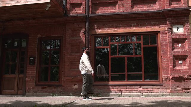 Adult man walking past weathered red brick building with large wooden framed windows, strong daylight shadows, urban sidewalk detail, and historic facade texture