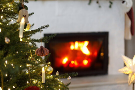 Close up of decorated Christmas tree with stylish ornaments and glowing lights on cozy burning fireplace background. Festive living room interior, winter holiday comfort.