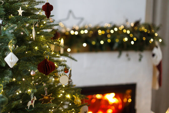 Close up of decorated Christmas tree with stylish ornaments and glowing lights on cozy burning fireplace background. Festive living room interior, winter holiday comfort.