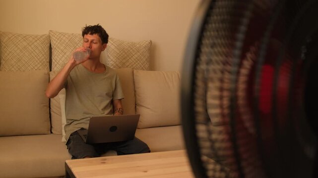 Man struggling with extreme heat at home, working on laptop and drinking water to stay productive during hot weather and high indoor temperature