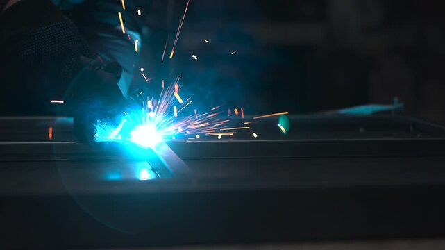 Bright welding arc with flying sparks in dark industrial workshop, worker silhouette leaning toward metal surface under intense blue white glow, strong contrast, reflective steel, gritty repair and