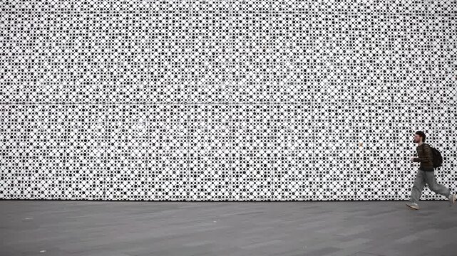 Pedestrian walking beside modern perforated metal facade beside modular panel grid and grey paved plaza, showing mid-stride movement, geometric texture, urban minimalism, and strong architectural