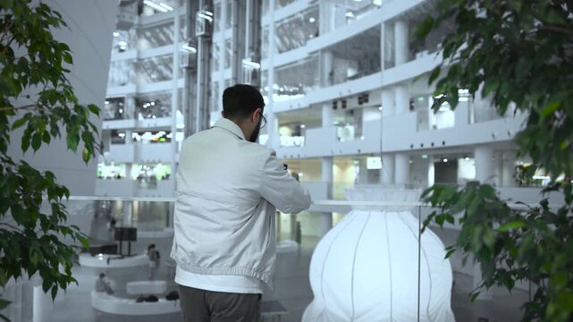 Adult man leaning on glass railing inside modern multi level atrium, using smartphone with leafy foreground framing, sculptural lamp below, and calm architectural interior light