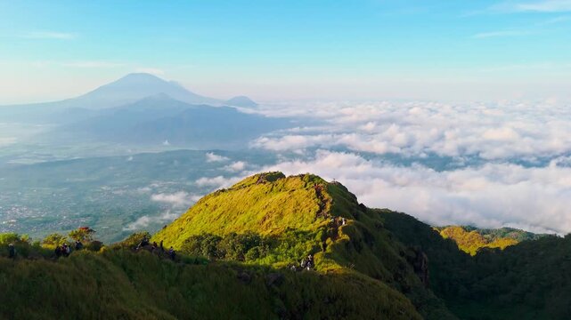 Aerial view of Mount Ungaran peak with hikers walking along the grassy ridge above a sea of clouds in Ungaran Barat, Central Java, Indonesia.