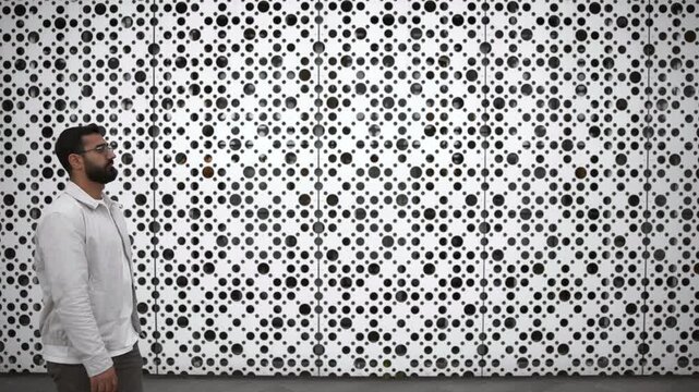 Bearded man walking beside large white perforated metal wall with circular hole pattern across expansive modern urban facade scene, with man, walking, bearded, profile detail