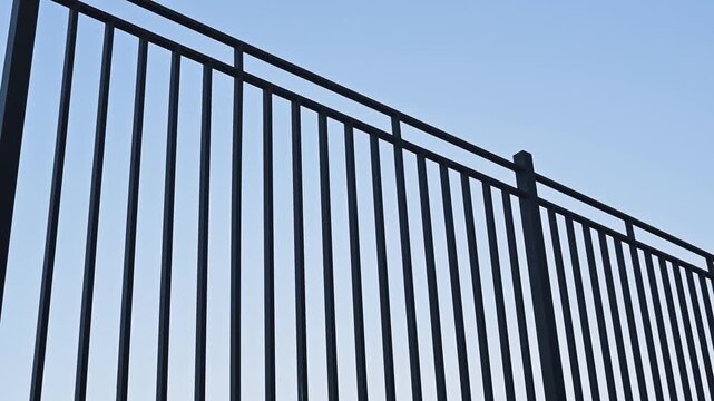 A black metal fence stands tall against a blue sky in a suburban area. The vertical bars of the fence create an organized pattern that offers structure and division of space