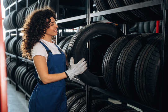 Female auto technician in blue overalls holds a tire while standing in a tire storage area with shelves filled with various tires in the background