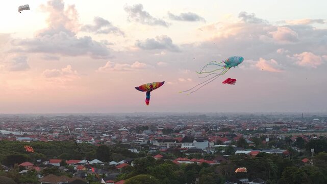 Aerial view of colorful kites flying above Mertasari Beach at sunset with the cityscape of Denpasar Selatan in the background, Denpasar Selatan, Bali, Indonesia.