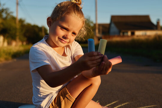 A young child sits on the pavement holding several pieces of chalk while smiling. The sun shines in the background on a late afternoon