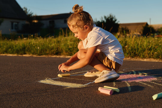 A young child crouches down and uses colorful chalk to draw on the pavement. The sun shines bright and the surroundings have green grass