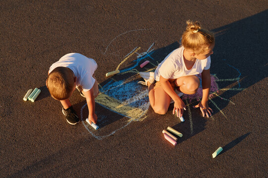 Two children are sitting on a grey pavement using chalk to create colorful drawings. It is sunny and they seem to enjoy their activity