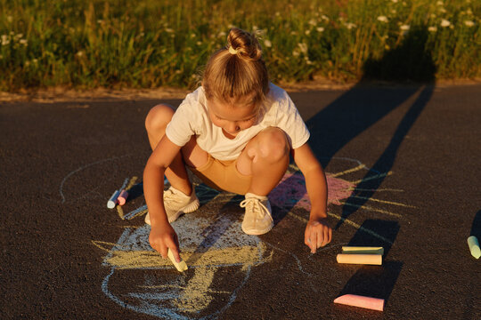 A child is sitting on a road drawing with colorful chalk while the sun sets. The grass around is green and the sky shows warm hues of a late afternoon