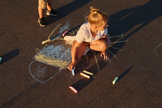 Two children play on the ground in a playground during a summer evening. One child sits and colors with chalk while the other stands nearby