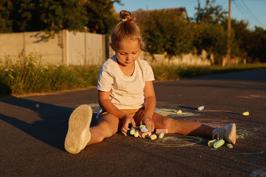 A child sits on the pavement and uses colored chalk to make drawings. Sunlight casts a warm glow, and trees are visible in the background