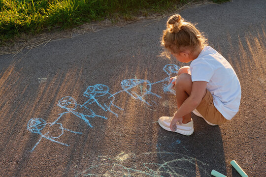 A young child sits on a sidewalk, using chalk to draw stick figures. The sun casts long shadows, and grass is visible nearby