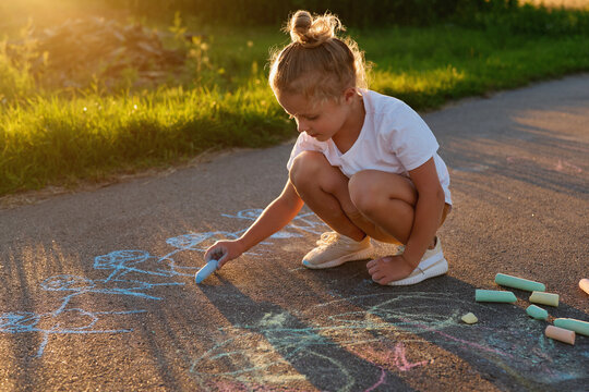 A young girl is sitting on the ground and using chalk to draw colorful pictures on the sidewalk as the sun sets behind her