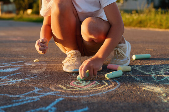 A child is crouched down on the pavement using colorful chalk to create drawings. The sun is setting, casting warm light in a residential area