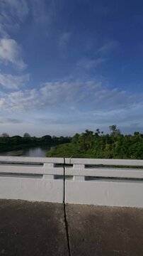 Scenic view from a bridge overlooking a calm river and lush tropical landscape beneath a blue sky with soft clouds, capturing peaceful nature, travel and countryside scenery.
