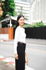 Fototapeta premium Urban fashion portrait of a young Asian woman standing by a city street in Bangkok, Thailand. She is looking at the camera, wearing a minimalist white long-sleeve top and black trousers with a soft bl