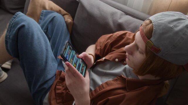 Caucasian teenager sitting on sofa and playing mobile game on smartphone. Perfect for marketing mobile gaming apps, digital entertainment, or screen time awareness campaigns