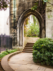Naklejka premium Beautiful medieval Gothic stone archway surrounded by lush greenery and a curved stone pathway in a historic English garden.