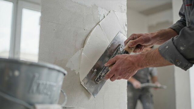 A professional construction worker uses a metal trowel to apply smooth plaster on a white wall during home renovation work