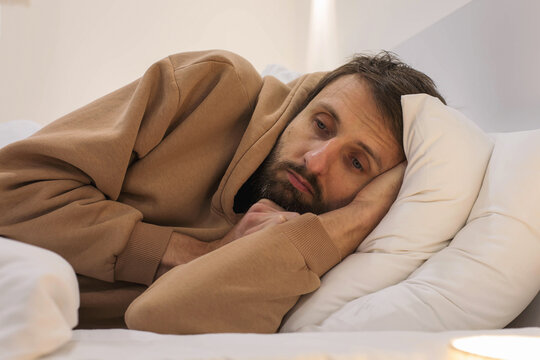 Exhausted bearded man lying awake in bed with sad expression, wearing casual hoodie, struggling with depression, burnout, emotional stress, insomnia, mental health, fatigue, recovery concept
