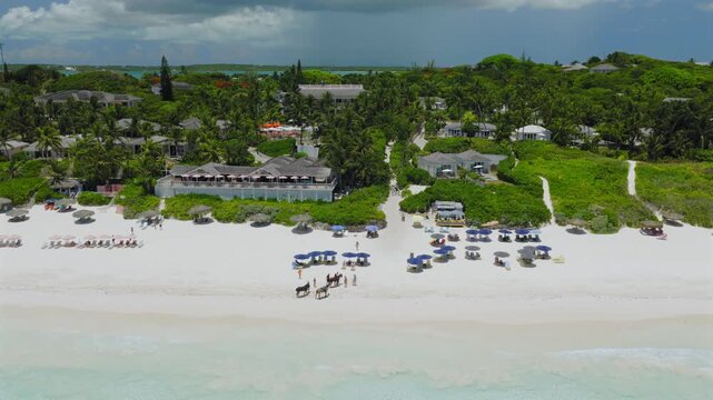 Aerial view of Pink Beach with resort buildings, umbrellas, and people riding horses on the sand in Rock Sound, South Eleuthera, The Bahamas.