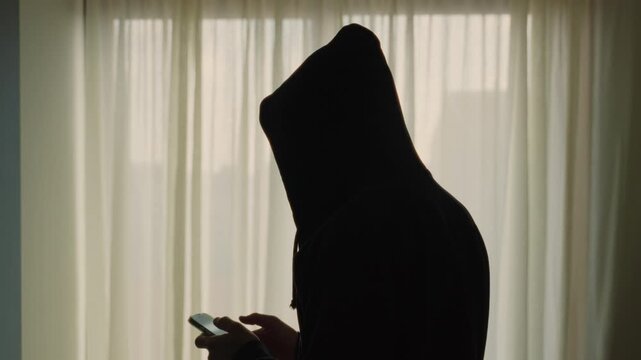 Mysterious hooded person's silhouette against sunny window curtain texting and using smartphone. Hacker or spy standing inside apartment or house holding mobile phone and writing message or chat