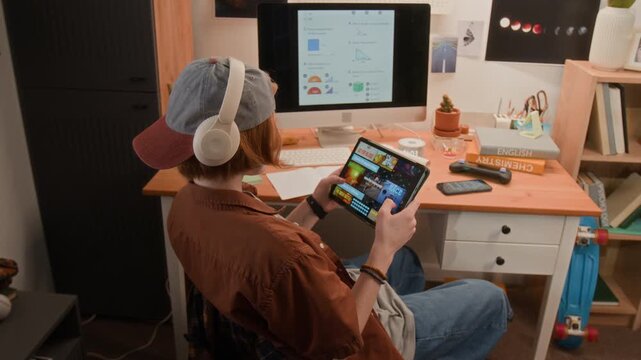 Caucasian teenager wearing headphones sitting at desk, using foldable tablet while computer screen displays charts. Video suitable for marketing e-learning apps and digital study tools