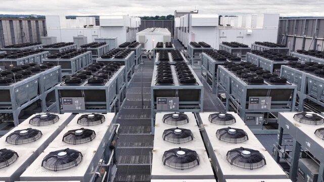 Rows of gray cooling units and industrial fans span data center rooftop in Clifton, New Jersey, narrow metal walkways separate dense equipment beneath overcast cloudy sky. Aerial drone view.