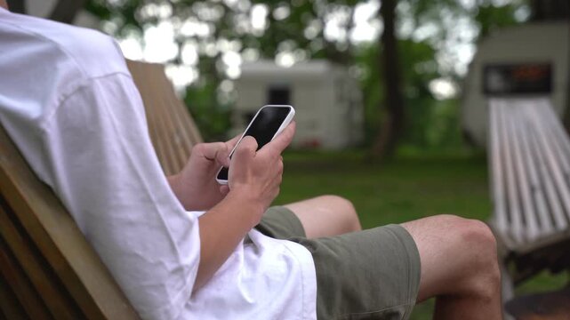 Person lounges in wooden chair, thumbs scrolling phone. Background shows green grass, trees, and another empty chair. Relaxation, vacation, or social media use themes. Outdoor leisure concept