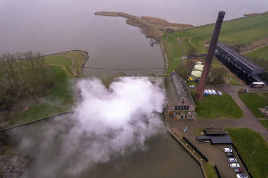 Aerial view of the historic Wouda pumping station with its tall chimney and steam venting over the water, surrounded by green dikes in Lemmer, Friesland, Netherlands.