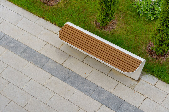Wooden park bench on a stone tile pavement next to a green lawn with shrubs. Modern outdoor street furniture in a landscaped residential courtyard area from a high angle view.