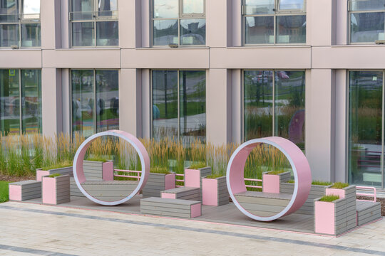 Pink circular street benches and wooden planters in front of a modern building facade. Contemporary urban furniture design and street art installation in a public courtyard area.