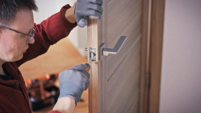 A profile shot of a technician wearing glasses and work gloves repairing a door lock on a grey brown wooden interior door with an orange screwdriver. Concept of home repair service