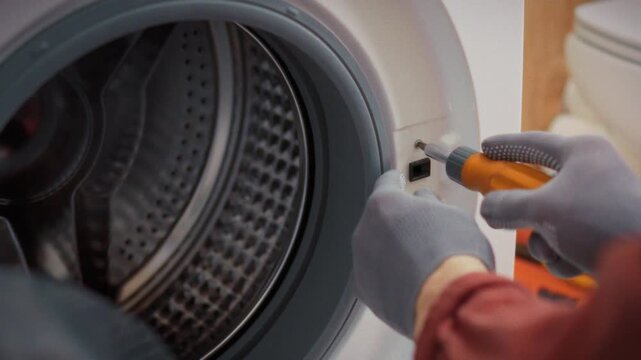 A specialist in work gloves disassembles a washing machine with a drum window unscrewing body bolts in a bathroom. Concept of scheduled technical maintenance of home appliances