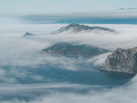 Aerial view of the Aleutian Islands near Unalaska, with dramatic fog rolling over rugged green mountains and rocky cliffs above the dark ocean, Unalaska, Alaska, United States.