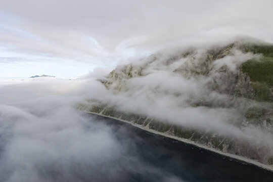 Aerial view of rugged coastal cliffs partially shrouded in thick low-hanging clouds above the dark waters of the Bering Sea in Unalaska, Alaska, United States.