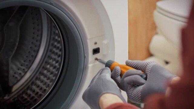 A technician in gloves methodically unscrews bolts from the washing machine body in a bathroom with a toilet and toolbox visible in the background. Concept of professional technical servicing