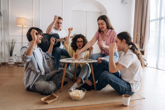 Group of young adults engaged in a board game night, sitting on the floor around a table with snacks, bright living room with large windows and modern decor