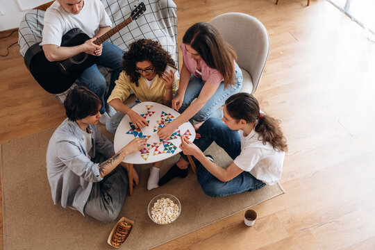 Group of young adults engaged in a board game activity around a table with snacks, guitar resting nearby, in a cozy indoor living room setting
