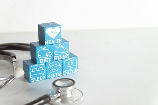 Healthcare and wellness concept. Blue wooden blocks with icons for health, diet, fitness, sleep, mental, and checkup stacked near a stethoscope on a white background with copy space.