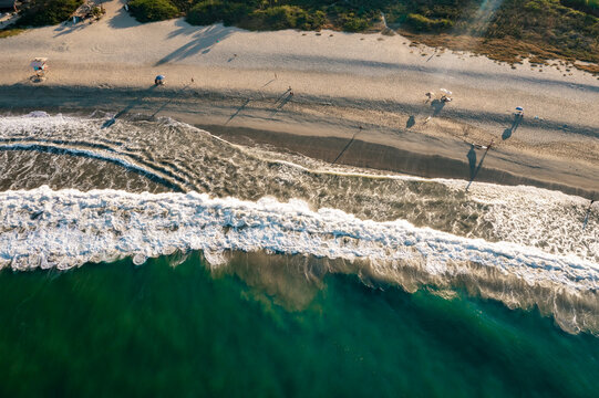 Aerial view of waves crashing onto the sandy beach with people and umbrellas under the warm golden hour light in Puerto Escondido, Oaxaca, Mexico.