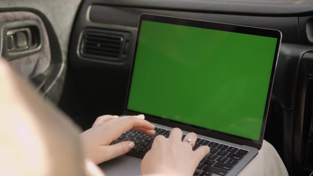 Person types on laptop with green screen inside car. Fingers move quickly over keyboard. Car interior visible, seatbelt nearby. Ideal for video editing or remote work concepts. Useful for advertising