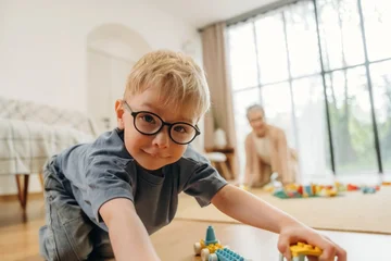 Fotobehang Lego Young boy with glasses playing with colorful building blocks on the floor in a bright living room, grandmother visible in the background engaging with toys  © standret