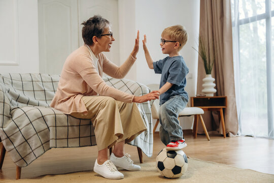 Elderly woman and young boy playing together at home, sitting on a sofa with a soccer ball, engaging in a joyful interaction in a bright living room setting