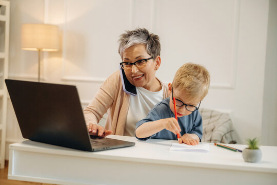 Elderly woman with short gray hair multitasking on a laptop while talking on the phone, young boy coloring beside her at a modern home office desk
